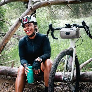Smiling black female cyclist sits on log with Fierce Hazel True Grit saddle bag on bike by her side.